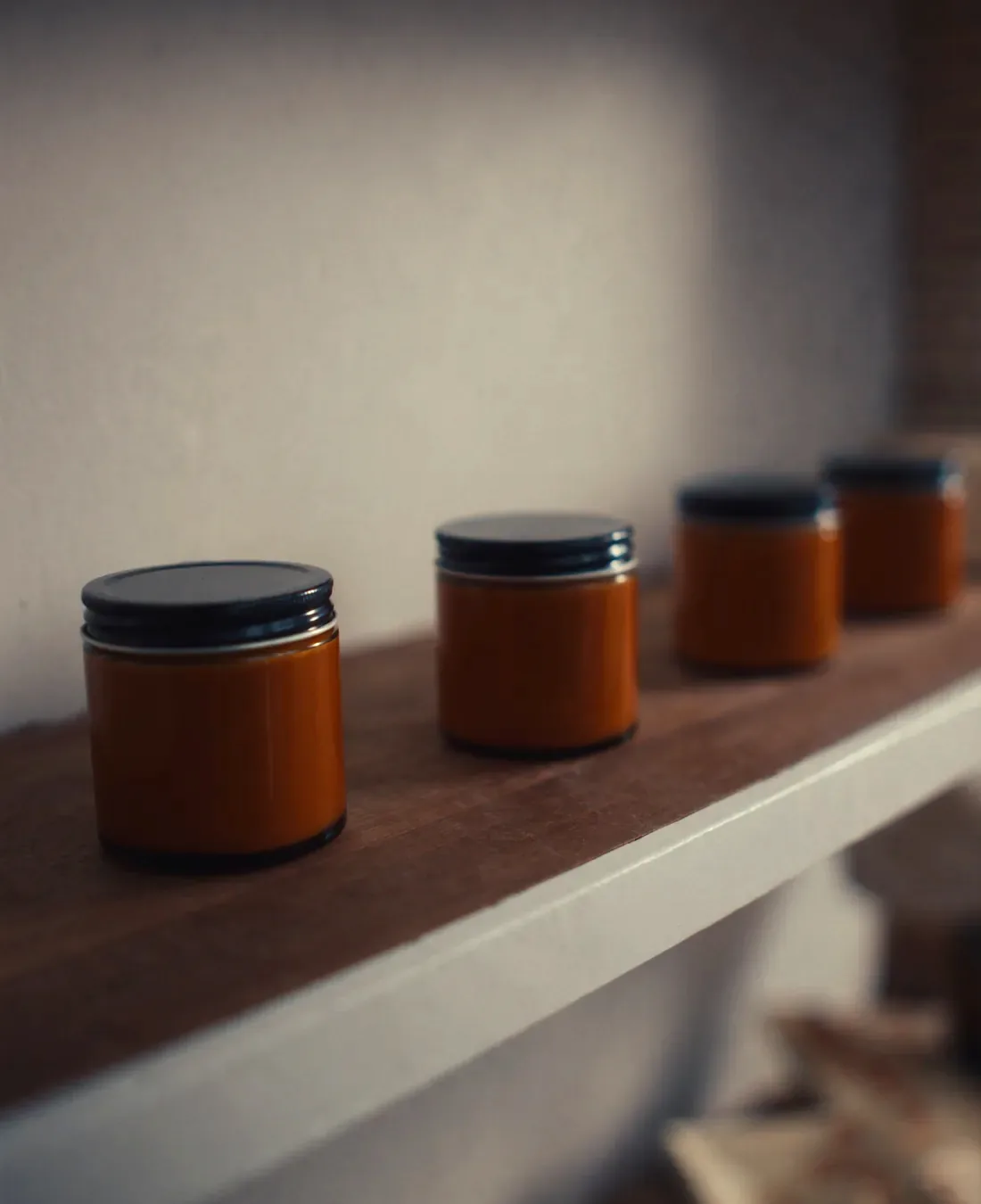 A shelf of poured soy candles in amber glass jars with plain lids on a painted wooden shelf, lit warmly from one side.