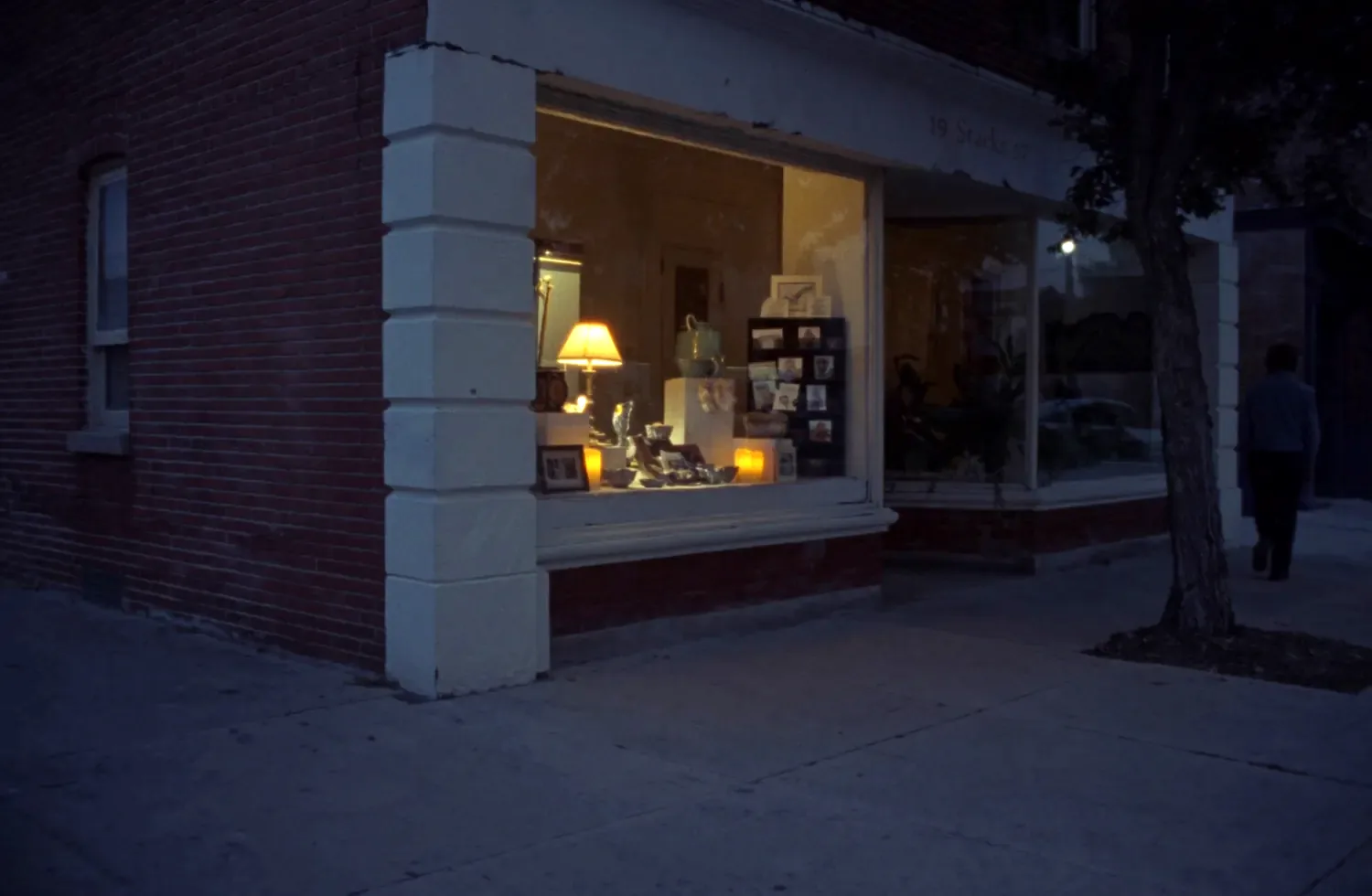 The lit display window of a small gift shop on the corner of a red brick building at dusk, with a lamp, small ceramics and a rack of cards visible behind the glass, and a figure walking away along the pavement.